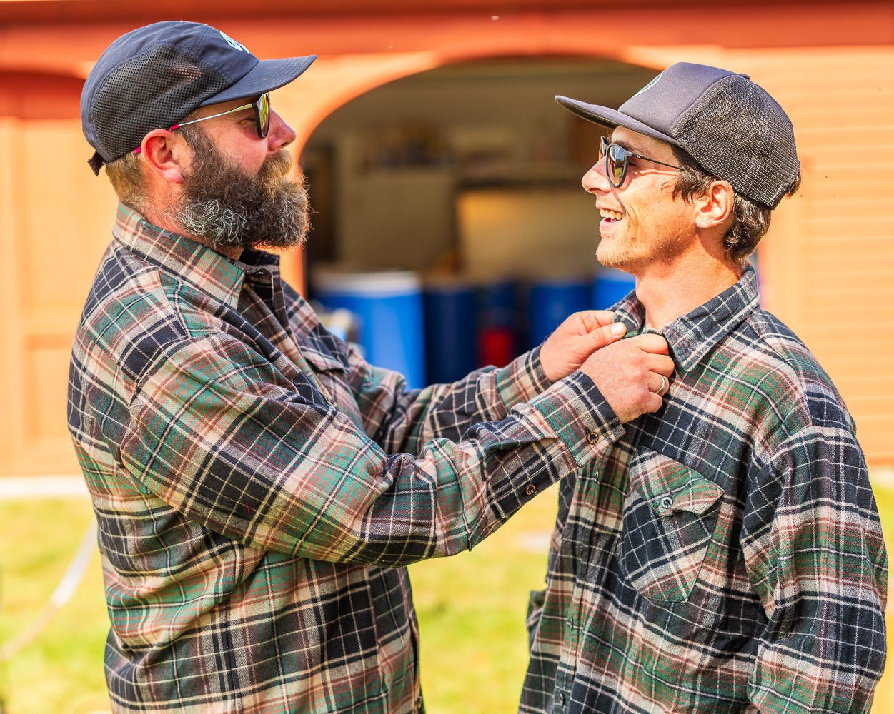 Two men wearing plaid shirts and caps standing in front of a barn.