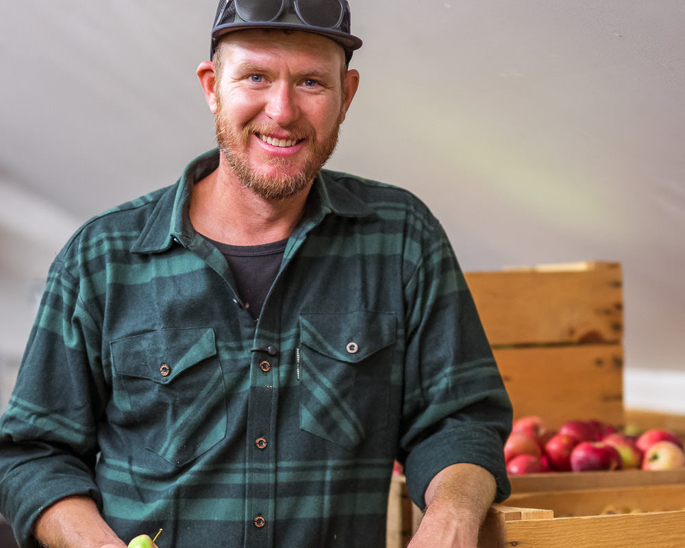 Man holding apples and standing next to wooden crates with apples inside, wearing a green plaid shirt and cap.